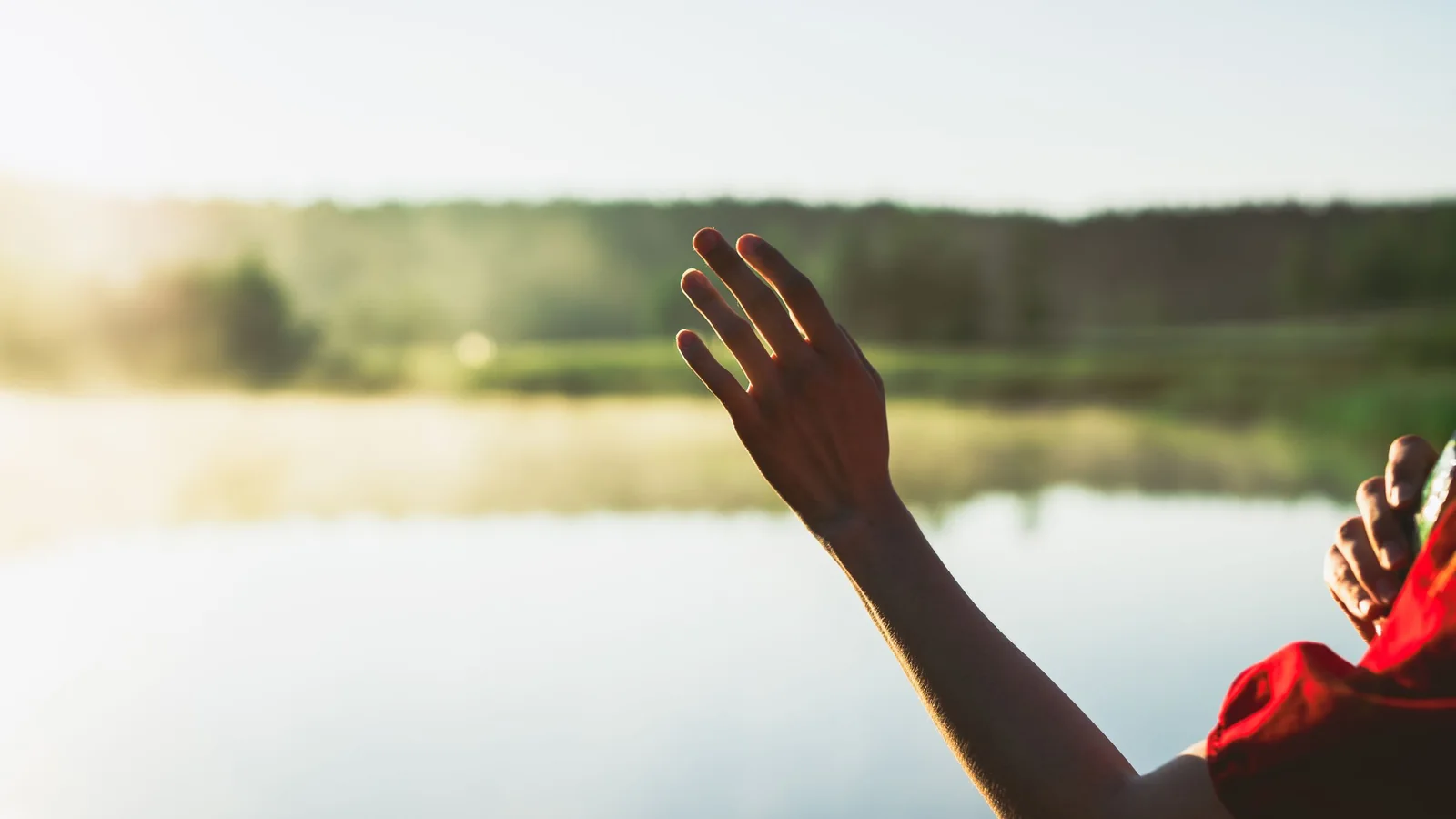 A woman raises one hand towards the light beside a calm lake.