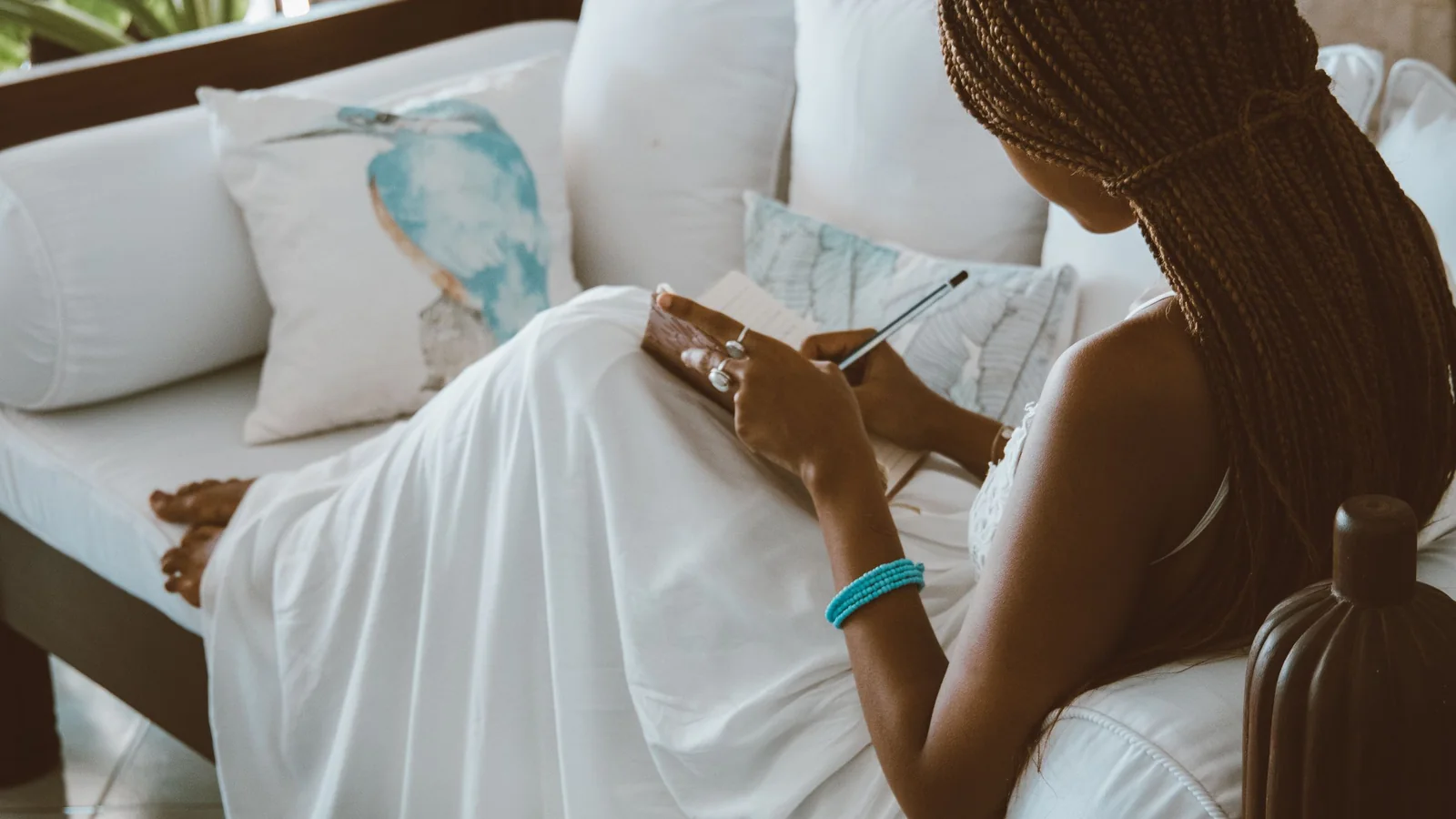 A woman sits on a bed writing in a notebook in soft daylight.
