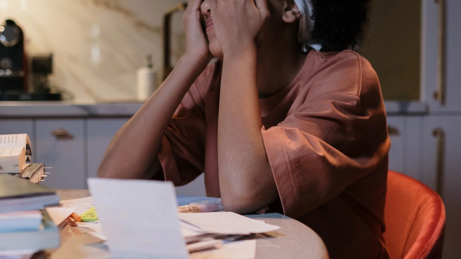 A tired woman sits at a kitchen table with her head resting in one hand.
