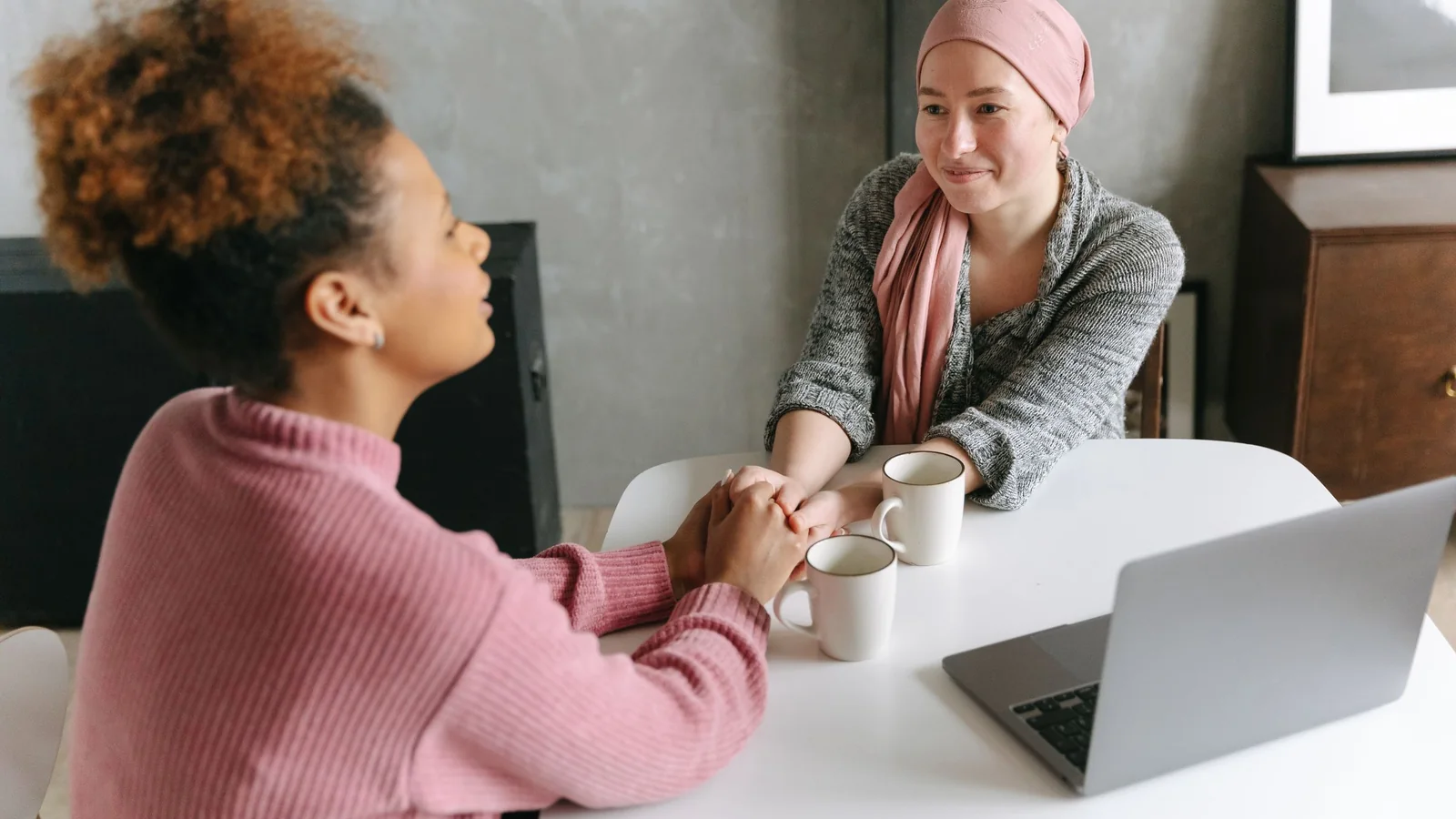 Two women sit at a table, holding hands as they talk.